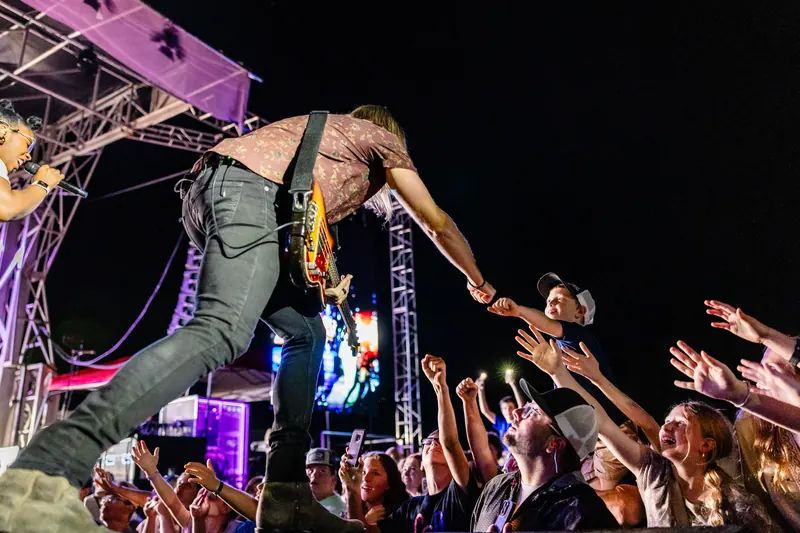 A guitarist leans off the edge of the stage at night to reach the outstretched hands of fans in the front row.