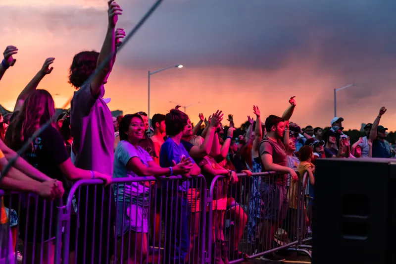 Fans at the front barricade raise their hands in worship under a pink and orange sunset sky.