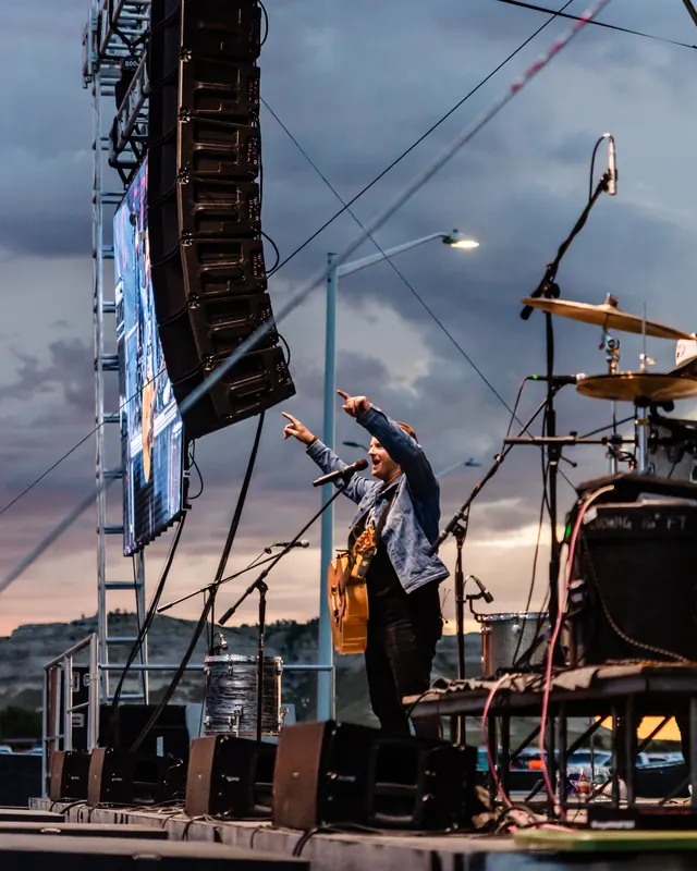 A singer performs on the main stage at sunset beside a tall speaker tower and drum kit.