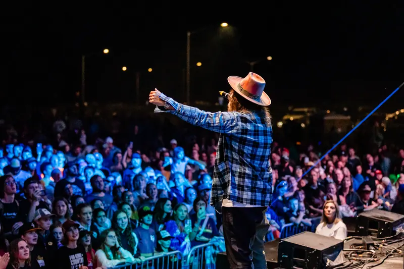 A performer in a wide brimmed hat and plaid shirt points out over a crowd lit in blue at night.