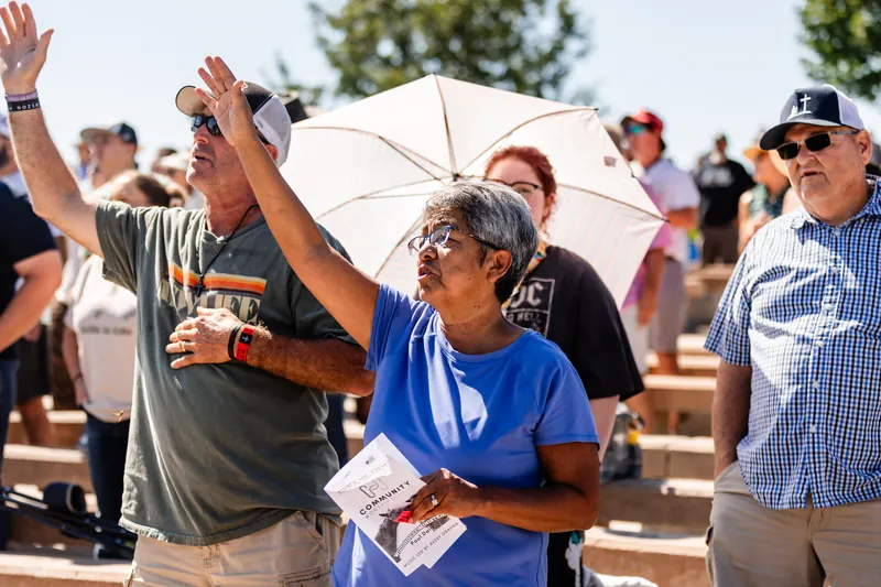 Two people in the outdoor amphitheater lift their hands in worship on a sunny morning.