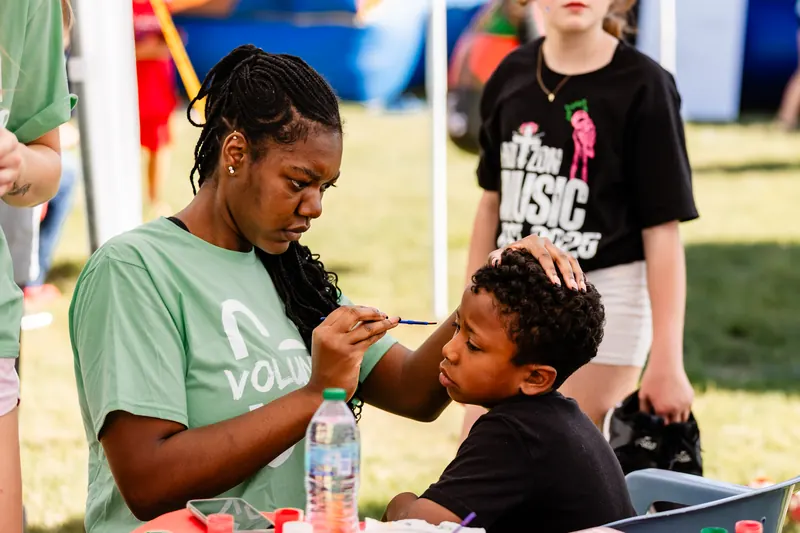 A volunteer in a green festival shirt paints a design on a young boy's face.