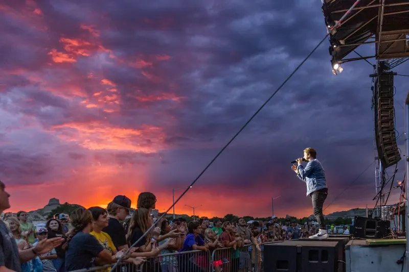 A singer stands at the front of the stage facing a large crowd beneath a dramatic red and purple sunset.