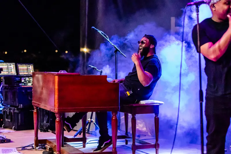 A musician smiles while playing a red organ on stage as blue light and haze fill the background.