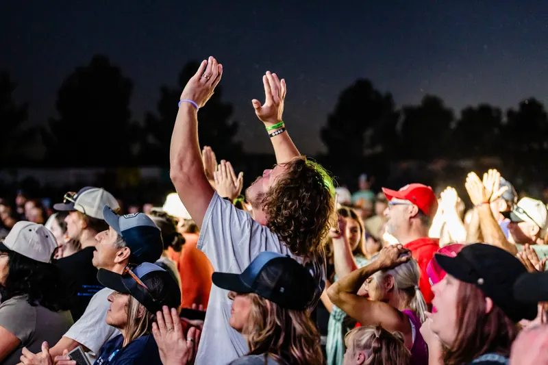 A crowd raises their hands toward the stage at dusk, with one man lifting both arms high.