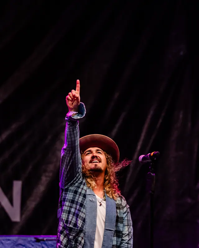 A singer in a hat and plaid shirt points one finger toward the sky during a night performance.