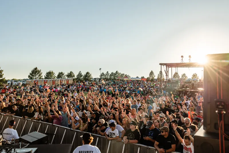 A large daytime crowd fills the field in front of the stage as the sun sits low behind it.
