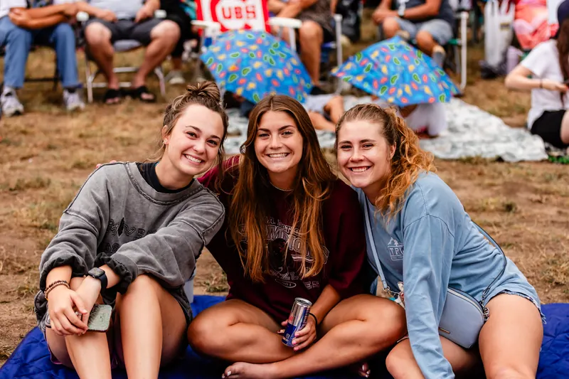 Three young women sit together on a blanket on the festival lawn and smile for the camera.