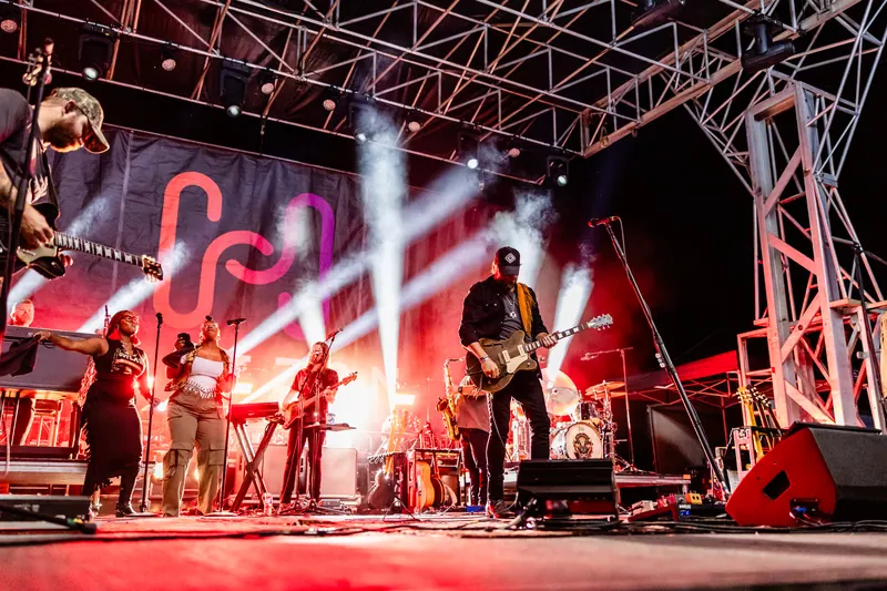 A full band performs under the lit Horizon stage truss with red lights and white spotlight beams.