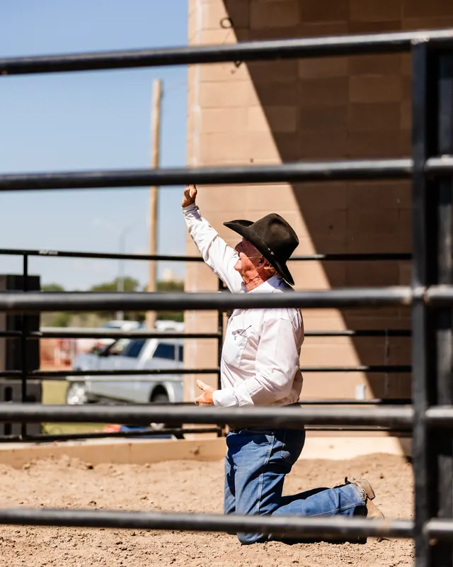 A man in a cowboy hat kneels in the dirt of the arena with one hand raised in prayer.