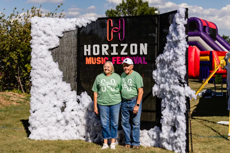Two volunteers in green shirts stand in front of a Horizon Music Festival photo wall.
