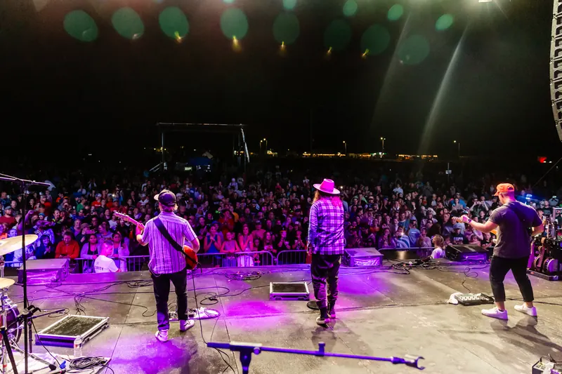 View from behind the band on stage looking out at a large crowd under purple stage lights.