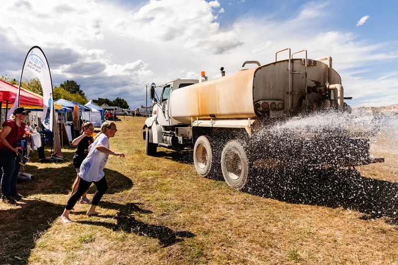 A water truck sprays the dry field on a sunny day as a child runs through the spray.