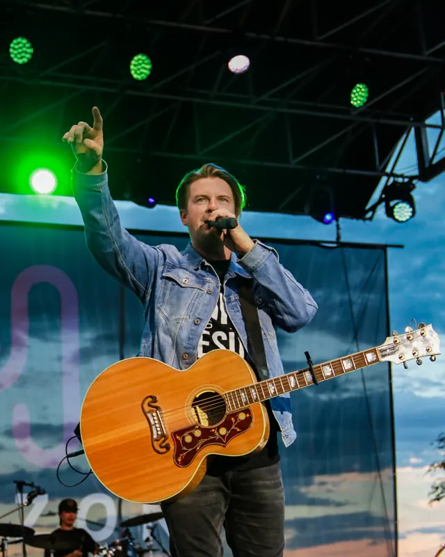 A singer with an acoustic guitar points to the crowd from the stage on a clear evening.