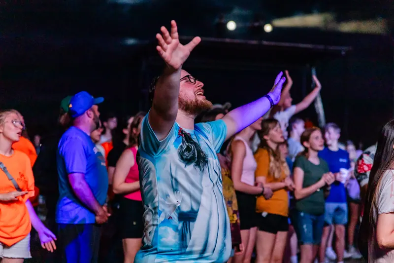 A man in a tie dye shirt lifts both hands in worship among a crowd at a night show.