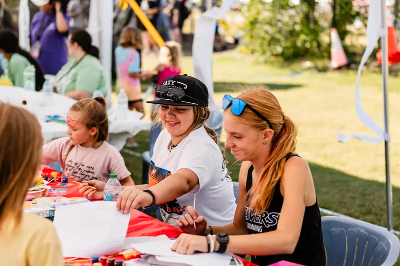 Kids and teens sit at a table working on arts and crafts in the shade of a tent.
