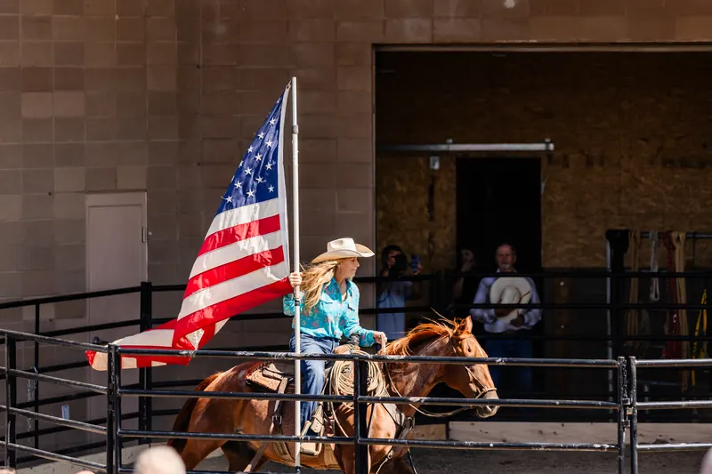 A rider in a cowboy hat carries an American flag on horseback in the arena.