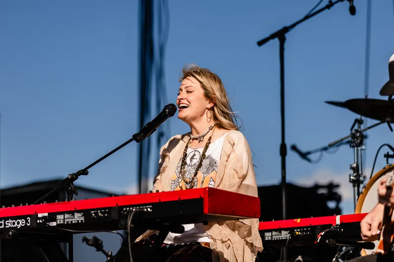 A singer plays keyboard and sings on stage under a bright blue daytime sky.