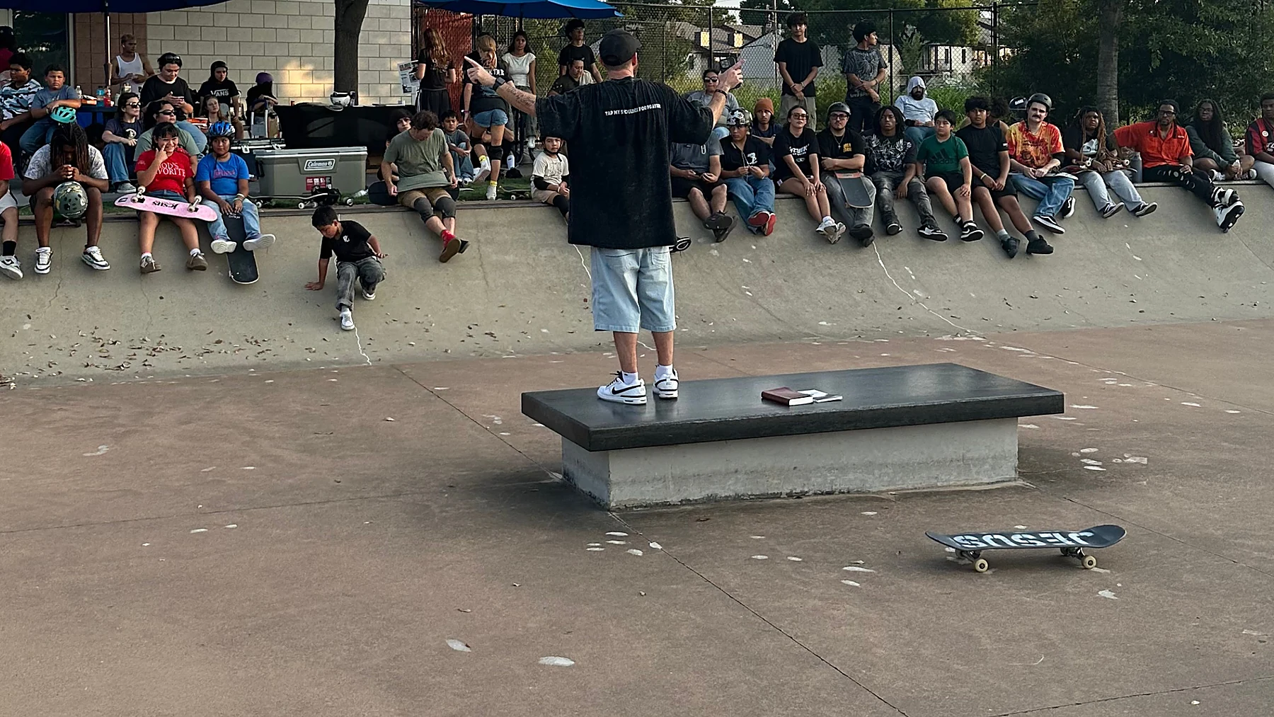 Kenny Hampton preaching from a skatepark ledge, his Bible open beside him, a crowd of skaters sitting along the coping