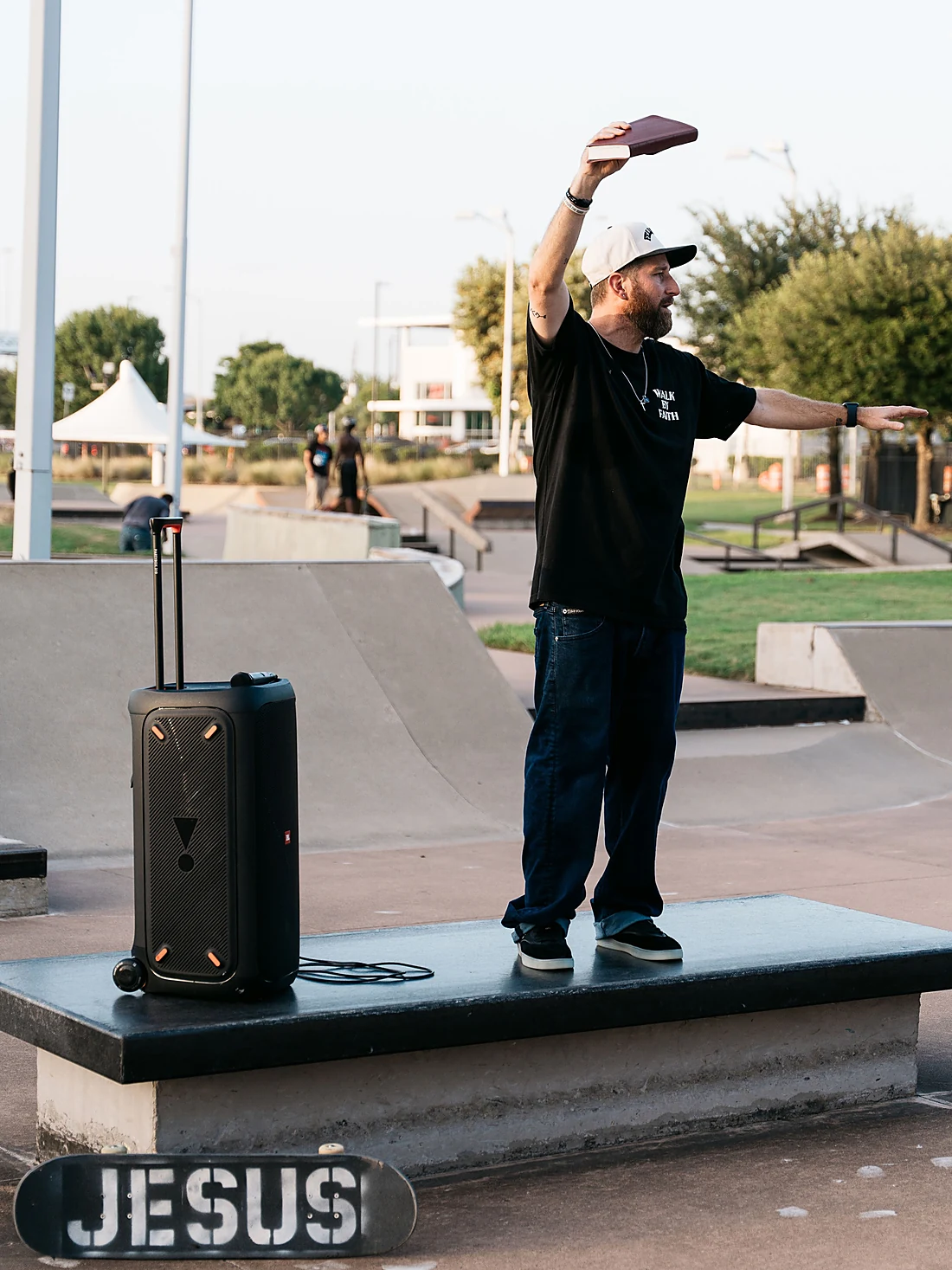 Kenny Hampton preaching at a skatepark with his Bible raised, a board reading JESUS on the ground in front of him